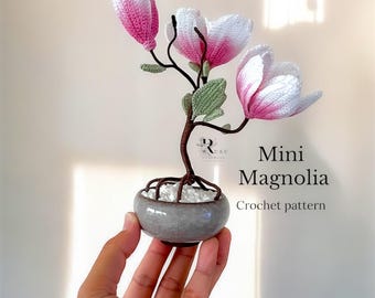 May include: A crocheted miniature magnolia tree in a gray pot. The tree has three pink and white flowers and green leaves. The text "Mini Magnolia Crochet pattern" is below the tree.