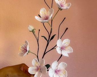 May include: A decorative branch featuring multiple white crocheted flowers with soft pink centers and bright yellow stamens. The branch has dark brown stems and small green buds, showcasing intricate needlework and detailed petal textures throughout.