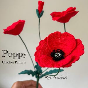 May include: A handmade crochet poppy flower arrangement. The arrangement features vibrant red poppies with black centers and green stems and leaves. The text "Poppy Crochet Pattern" is visible in the image.