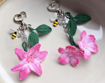 May include: Two handmade keychains featuring pink crocheted flowers with green leaves and a silver bee charm. The keychains have silver heart-shaped clasps. The flowers have a gradient of pink and white.