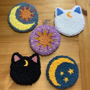 May include: Five round, colorful, embroidered felt coasters with moon and star designs. The coasters feature a yellow moon, a white cat, a purple sun, a black cat, and a blue moon with stars.