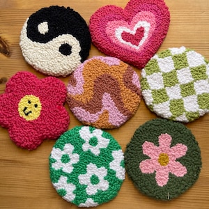May include: Seven colorful round punch needle wall hangings. The designs include a yin yang symbol, a heart with a smaller heart inside, a swirl pattern, a checkered pattern, a flower with a smiley face, a green background with white flowers, and a green background with a pink flower.