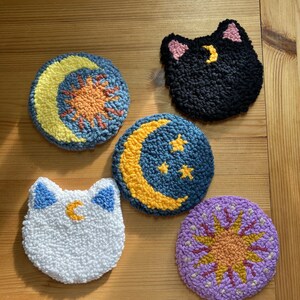 May include: Five handmade needle felted ornaments with celestial themes. The ornaments are round and feature a moon, sun, and stars in various colors. Two of the ornaments are shaped like cats with a crescent moon on their foreheads.