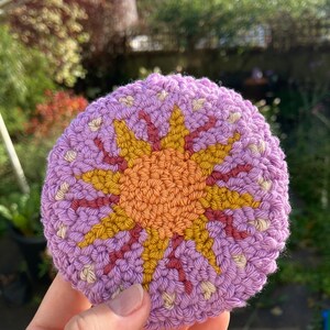 May include: A crocheted sun with a purple background, orange center, and yellow rays. The sun is crocheted with different colored yarns.