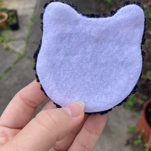 K&ouml;nnte beinhalten: Schwarze und wei&szlig;e geh&auml;kelte Katzenkopf-Applikation mit wei&szlig;em Filzr&uuml;cken.