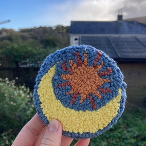 May include: A blue, yellow, and orange crocheted sun and moon design. The sun is in the center and the moon is a crescent shape around the sun.