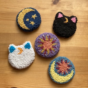 May include: Five round, colorful, embroidered felt ornaments with different celestial designs. The designs include a crescent moon with stars, a black cat with a crescent moon, a sun with rays, a white cat with a crescent moon, and a crescent moon with a sun.