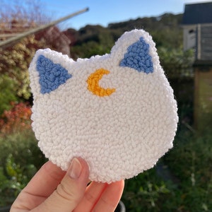May include: White crocheted cat head with blue ears and a yellow crescent moon embroidered on the forehead.