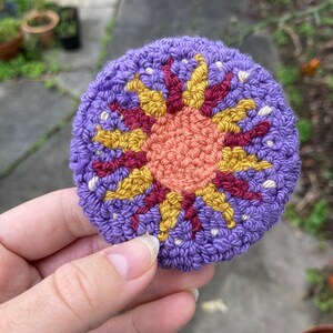 K&ouml;nnte beinhalten: Ein lila geh&auml;kelter Kreis mit einem bunten gestickten Sonnendesign. Die Sonne hat eine rote, orange und gelbe Mitte mit gelben Strahlen.