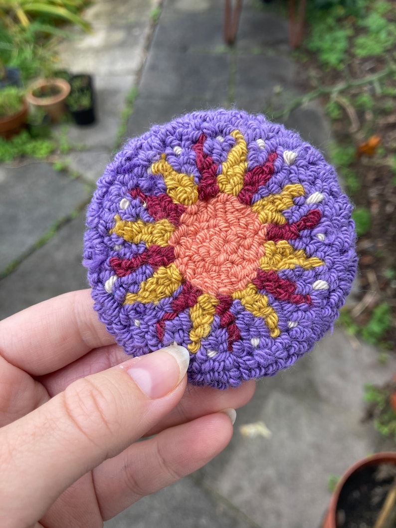 K&ouml;nnte beinhalten: Ein lila geh&auml;kelter Kreis mit einem bunten gestickten Sonnendesign. Die Sonne hat eine rote, orange und gelbe Mitte mit gelben Strahlen.