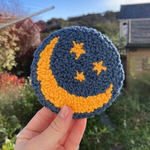 May include: A blue and yellow crocheted moon and stars ornament. The moon is a crescent shape and is outlined in yellow yarn. There are three yellow stars on the blue background.