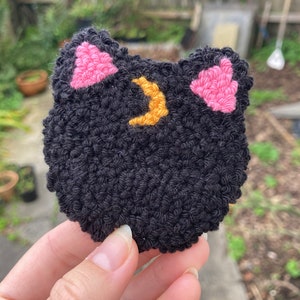 K&ouml;nnte beinhalten: Schwarzer Filzkatzenkopf mit rosa Ohren und einem gelben Halbmond auf der Stirn.