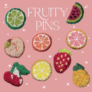 May include: A set of nine colourful felt fruit pins, including a lime, watermelon slice, peach, orange slice, lemon slice, strawberry, pineapple, and two cherries. The pins are arranged on a pink background with the text "FRUITY PINS" in white.
