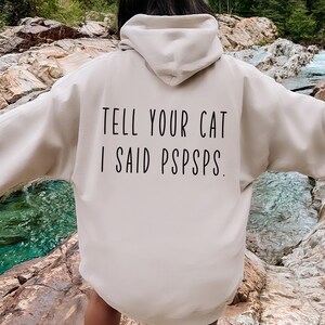 Puede incluir: Una sudadera con capucha beige con el texto "TELL YOUR CAT I SAID PSPSPS." impreso en la espalda.
