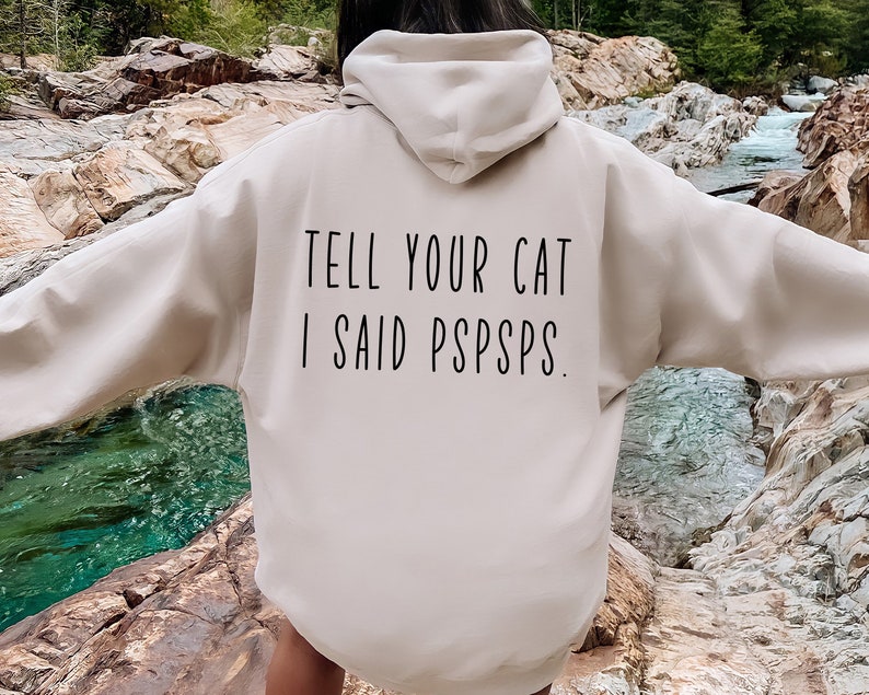 Puede incluir: Una sudadera con capucha beige con el texto "TELL YOUR CAT I SAID PSPSPS." impreso en la espalda.