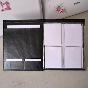 Könnte beinhalten: Offenes schwarzes Lederportfolio mit Taschen und Klarsichthüllen mit weißen Karten. Auf der linken Seite befindet sich ein Aufkleber mit einer Nähmaschine, und im Hintergrund ein Brother-Drucker mit floralen Akzenten.