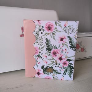 May include: A journal with a floral pattern and a pink scalloped edge. The cover displays pink roses, green leaves, and a small wooden tag. The journal is set against a white background.
