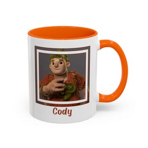May include: White ceramic mug with an orange rim and handle. The mug features a cartoon character with brown hair and a red shirt. The character is holding a green object. The mug has the word "Cody" printed on the bottom.