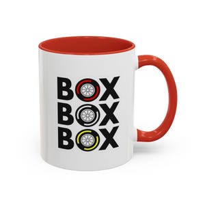 May include: White ceramic mug with a red interior and handle. The mug features the word "BOX" stacked three times, each with a different colored tire graphic: red, silver, and yellow. A fun, racing-themed design.