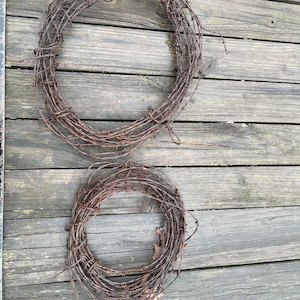 May include: Two circular wreaths made of rusty barbed wire. The wreaths are arranged on a weathered wooden surface. The barbed wire has a dark brown color, and the wood has a gray tone.