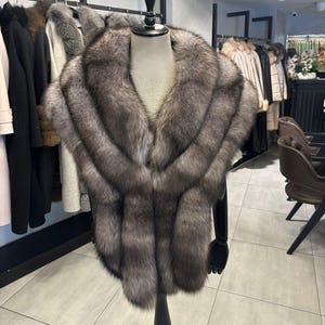 May include: A grey fur stole, displayed on a black mannequin. The stole features a layered design and a deep V-neck. Other fur coats and jackets are visible in the background, suggesting a high-end fashion boutique.