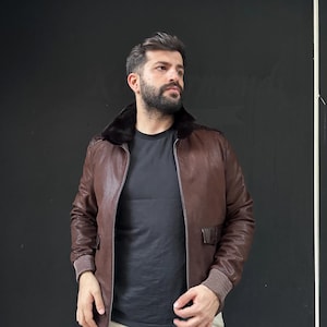 Könnte beinhalten: Braune Leder-Bomberjacke mit schwarzem Pelzkragen. Die Jacke hat gerippte Bündchen und ein strukturiertes Muster. Sie verfügt über zwei Vordertaschen und einen Reißverschluss. Die Jacke ist ein klassischer Stil.