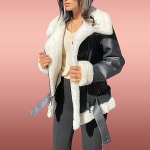 以下が含まれることがあります： 白いフェイクファーの襟と裏地が付いた黒いフェイクレザーのジャケット。ジャケットはジッパー留め、ベルト、複数のポケットが付いています。
