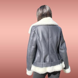 以下が含まれることがあります： 白いシープスキン襟とトリムが付いた黒い革のジャケット。ジャケットはウエストにベルトが付いており、白いシープスキンで裏打ちされています。