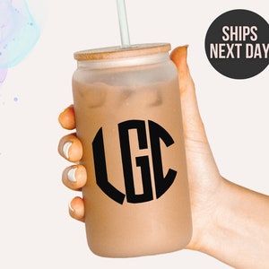 以下が含まれることがあります： 竹製の蓋と「LGC」と書かれた黒いモノグラムステッカーが付いた、フロスト加工されたガラス製のタンブラー。タンブラーは茶色の液体で満たされ、ストローが付いています。