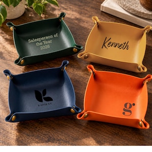 May include: Four square valet trays made of faux leather in dark green, yellow, tan, and orange. Each tray features gold-tone metal snaps at the corners and custom black text or logos, including Salesperson of the Year 2026, Kenneth, KAPIRNY, and a g.