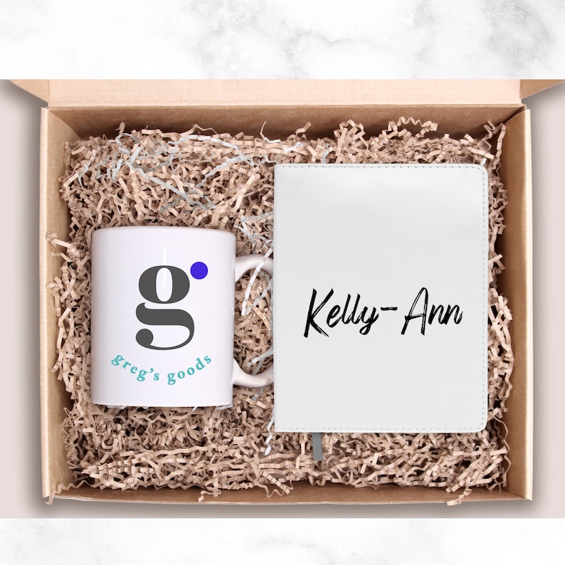 May include: A gift box filled with brown packing paper contains a white mug with a blue dot and the text "greg's goods" and a white journal with the text "Kelly-Ann" in black.