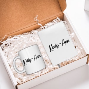 May include: A white gift box filled with white shredded paper. Inside the box is a white mug and a white notebook. Both items have the name "Kelly-Ann" written in black cursive.