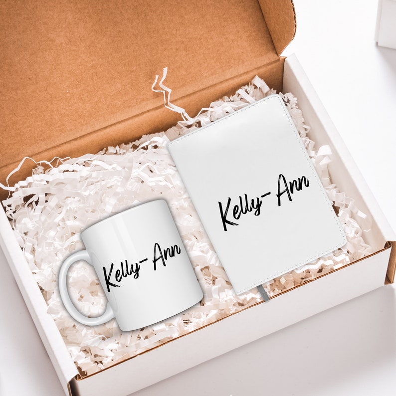 May include: A white gift box filled with white shredded paper. Inside the box is a white mug and a white notebook. Both items have the name "Kelly-Ann" written in black cursive.