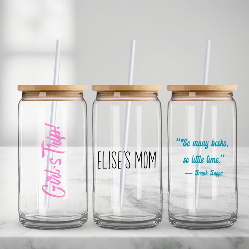 May include: Three clear glass canisters with bamboo lids and white straws. The canisters have different text printed on them: "Girl's Trip!" in pink, "ELISE'S MOM" in black, and a quote by Frank Zappa in blue: "So many books, so little time."