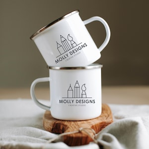 May include: Two white enamel mugs stacked on top of each other. Each mug has a silver rim and a white handle. The mugs feature a black logo with the text "MOLLY DESIGNS CREATIVE STUDIO" and a graphic of a pencil, paintbrush, and building.