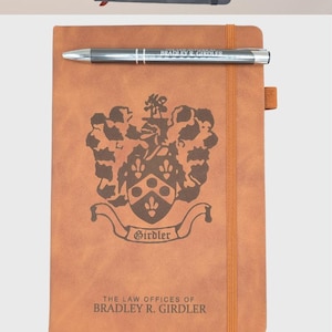 May include: Three personalized leather-bound journals with different designs. The first journal is gray with a geometric logo and the text "YOUR LOGO HERE". The second journal is brown with a crest and the text "THE LAW OFFICES OF BRADLEY R. GIRDLER". The third journal is blue with the text "KRISTIN".