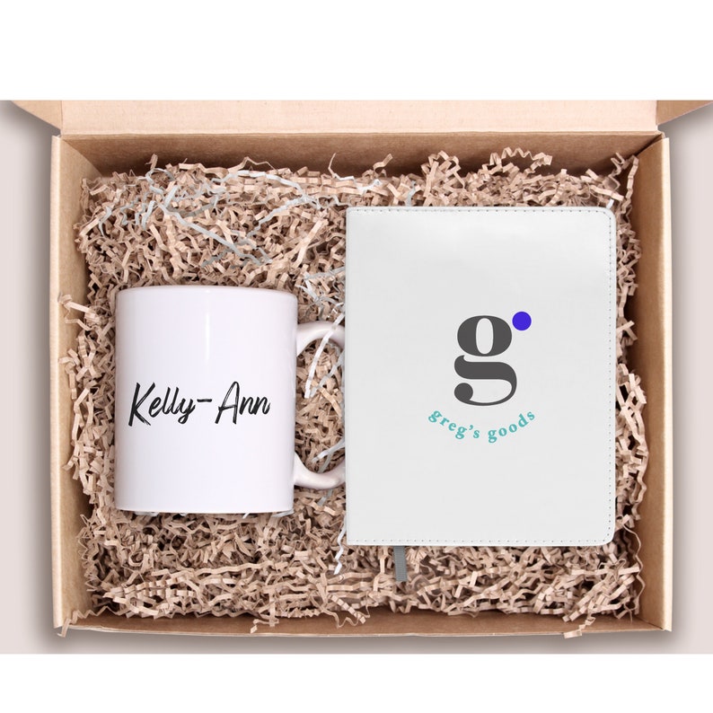 May include: A white mug with the name "Kelly-Ann" printed on it and a white notebook with the text "g. greg's goods" and a blue dot. The items are in a brown box with shredded paper.