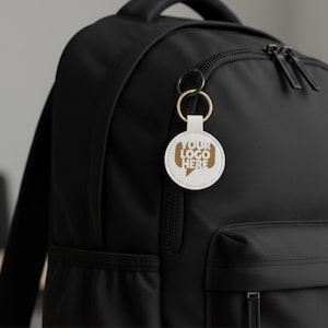 Puede incluir: Un llavero blanco y redondo con el texto "YOUR LOGO HERE" en un dise&ntilde;o de bocadillo marr&oacute;n, sujeto a un anillo dorado. El llavero cuelga de una mochila negra.