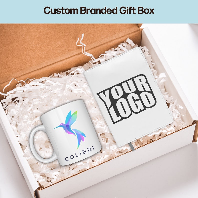 May include: A white gift box filled with white tissue paper. The box contains a white mug with a colorful hummingbird and the word "COLIBRI" printed on it. There is also a white notebook with the text "YOUR LOGO" printed in black.