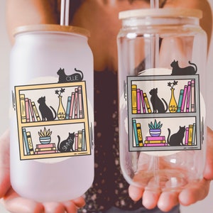 May include: Two clear glass tumblers with lids and straws. The tumblers feature a graphic of a bookshelf with cats and books. One tumbler has a yellow bookshelf and the other has a grey bookshelf. The text "Ollie" and "Sammy" are on the tumblers.