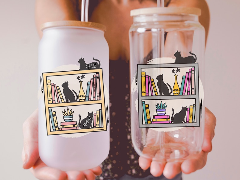 May include: Two clear glass tumblers with lids and straws. The tumblers feature a graphic of a bookshelf with cats and books. One tumbler has a yellow bookshelf and the other has a grey bookshelf. The text "Ollie" and "Sammy" are on the tumblers.