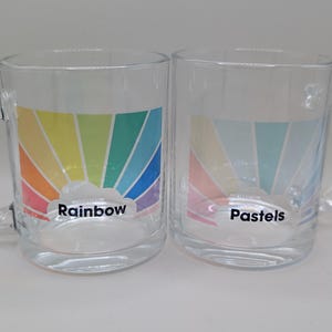 May include: Two glass mugs with rainbow designs. The mug on the left has a rainbow with a cloud and the text "Rainbow". The mug on the right has a pastel rainbow with a cloud and the text "Pastels".