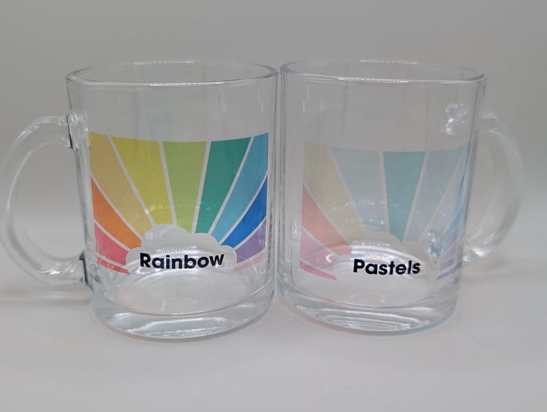 May include: Two glass mugs with rainbow designs. The mug on the left has a rainbow with a cloud and the text "Rainbow". The mug on the right has a pastel rainbow with a cloud and the text "Pastels".