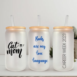 May include: Three frosted glass tumblers with bamboo lids and clear straws. The tumblers have different text printed on them: "Cat Mom", "Books are my love language", and "Career Week 2023".
