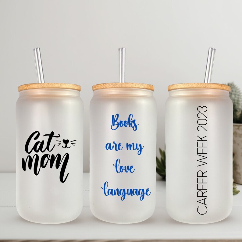May include: Three frosted glass tumblers with bamboo lids and clear straws. The tumblers have different text printed on them: "Cat Mom", "Books are my love language", and "Career Week 2023".