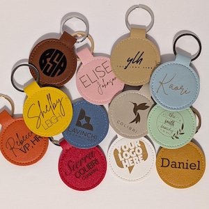 Puede incluir: Una colecci&oacute;n de llaveros personalizados en varios colores, incluyendo marr&oacute;n, rosa, amarillo, azul, rojo, beige, blanco y verde. Cada llavero presenta texto o logotipos personalizados, con anillos plateados. Los llaveros son redondos y est&aacute;n hechos de cuero o material similar.