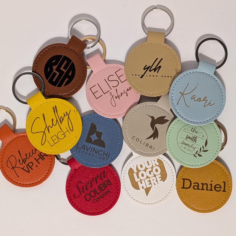 Puede incluir: Una colecci&oacute;n de llaveros personalizados en varios colores, incluyendo marr&oacute;n, rosa, amarillo, azul, rojo, beige, blanco y verde. Cada llavero presenta texto o logotipos personalizados, con anillos plateados. Los llaveros son redondos y est&aacute;n hechos de cuero o material similar.