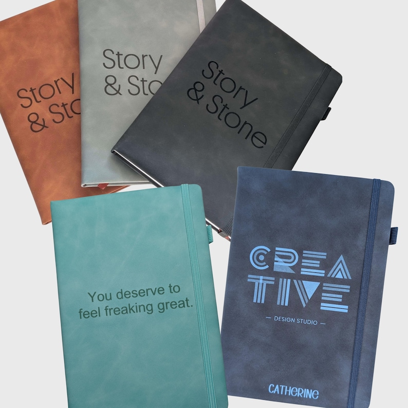 May include: Five rectangular notebooks with elastic closures and pen loops. Colors include tan, light gray, black, teal, and navy blue. The covers feature text: Story & Stone, You deserve to feel freaking great, and CREATIVE DESIGN STUDIO CATHERINE.