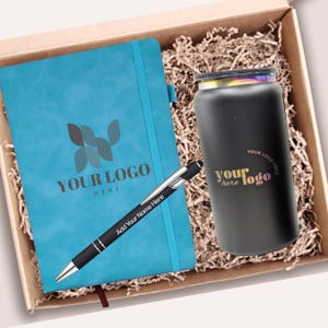 Pode incluir: Uma caixa de presente contendo um caderno azul-petróleo com um logotipo e o texto "YOUR LOGO HERE", uma caneta preta com "Add Your Name Here" e um copo preto com um logotipo. A caixa é preenchida com papel picado.