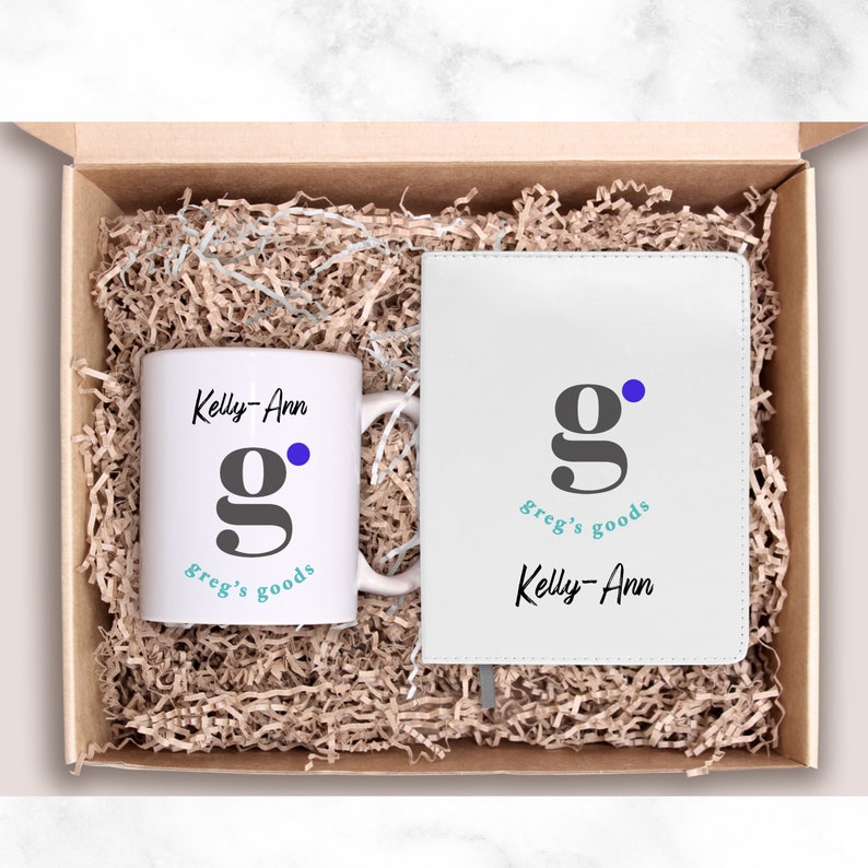 May include: A white gift box filled with brown packing peanuts. The box contains a white mug with a gray "g" and blue dot, and a white notebook with a gray "g" and blue dot. The mug and notebook both have the text "greg's goods" and "Kelly-Ann" printed on them.